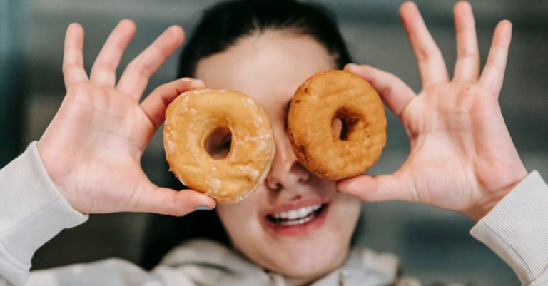 Portion Sizes - Positive young female in casual clothes smiling while covering eyes with delicious doughnuts